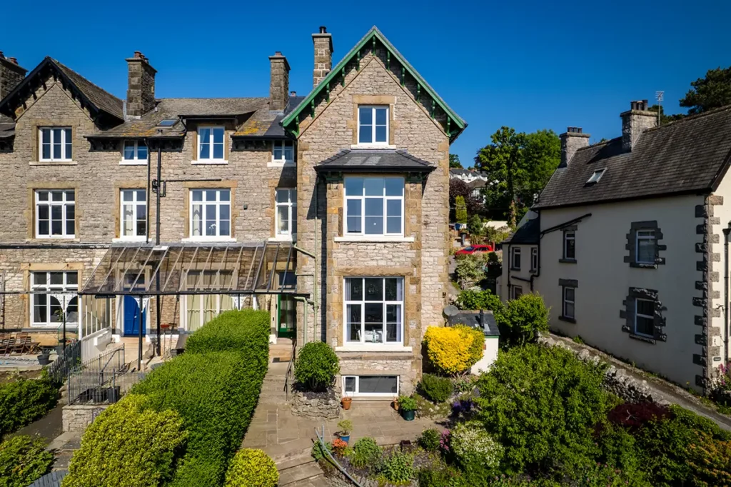 A large stone townhouse with tall windows, green trim on the gable, and a well-kept garden, set between neighboring houses under a clear blue sky.